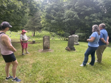 Two graves are centered in this photo with Dr. Natalie Woodall wearing a pink hat and reading notes. Three visitors stand and listen.
