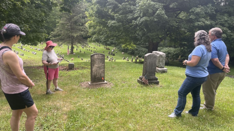 Two graves are centered in this photo with Dr. Natalie Woodall wearing a pink hat and reading notes. Three visitors stand and listen.