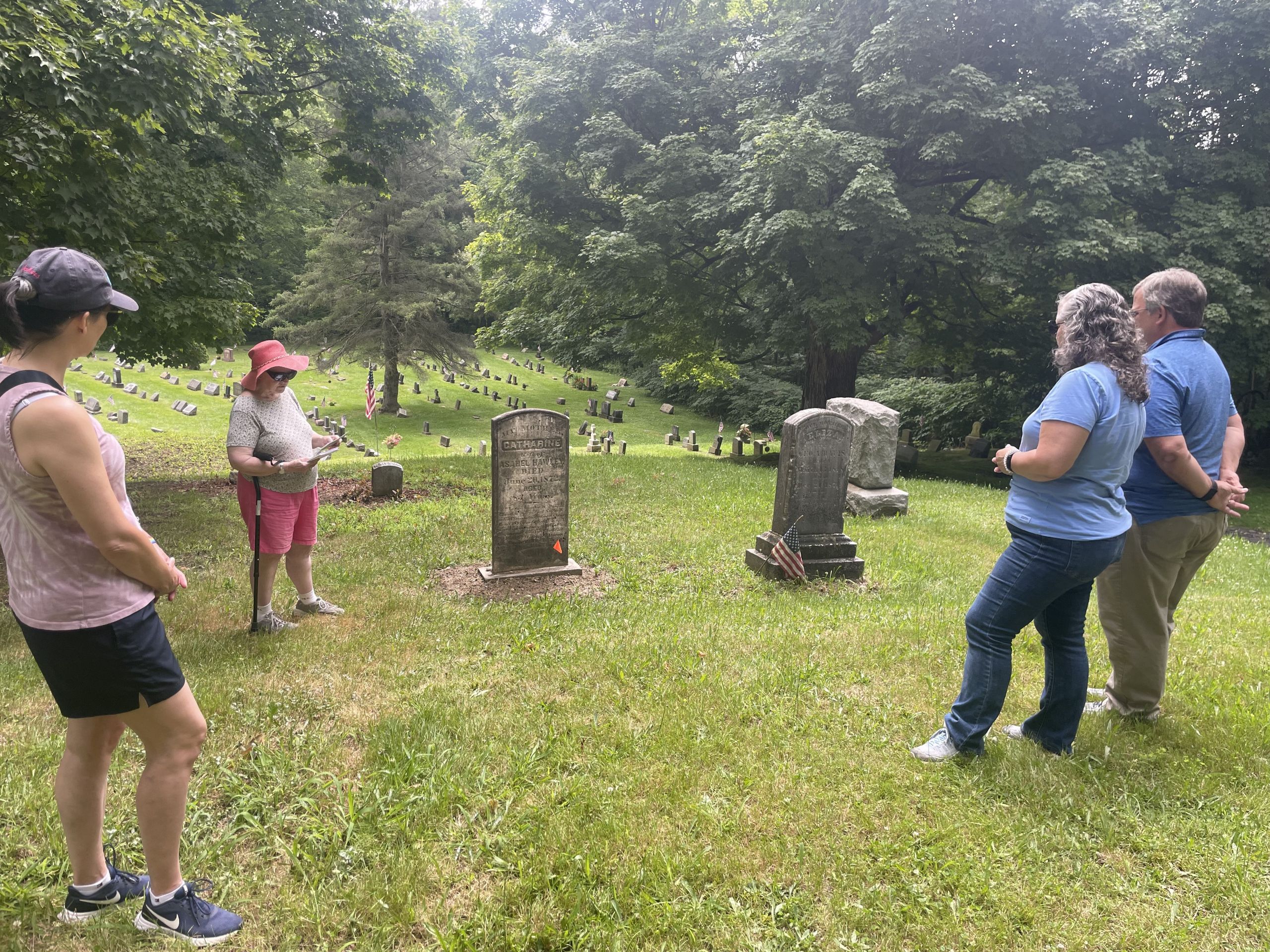 Two graves are centered in this photo with Dr. Natalie Woodall wearing a pink hat and reading notes. Three visitors stand and listen.